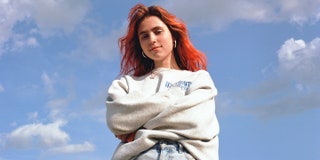Clairo has orange-dyed hair and is wearing gold hoop earrings, a grey sweatshirt, a gold necklace, and blue jeans. She's standing with her arms crossed, and a cloudy blue sky is the photo's background. 